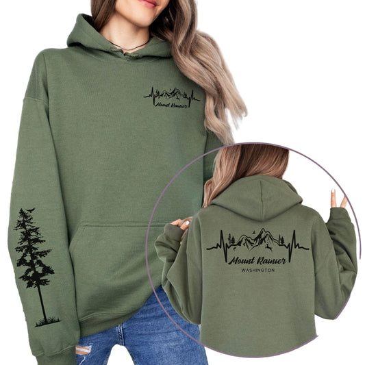 A green hoodie with 'Mount Rainier' and a heartbeat line design, featuring a pine tree on the sleeve. The hoodie is worn by a woman in a field with mountains in the background.
