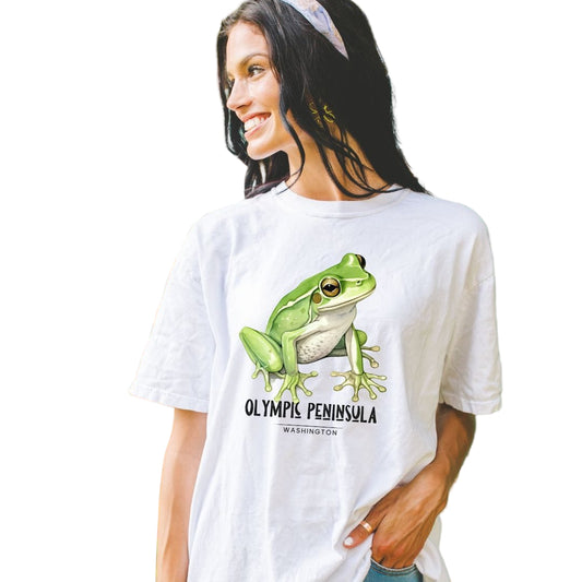 A woman wearing a white vintage-style t-shirt with a green frog graphic and the text 'Olympic Peninsula' on it, standing outdoors.